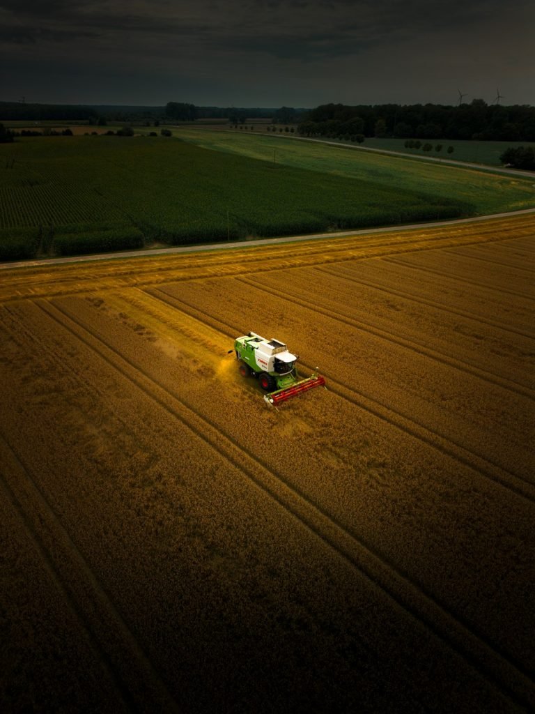 Aerial view of a harvester working in a golden wheat field, with surrounding green fields and distant wind turbines under a cloudy sky in Münster.